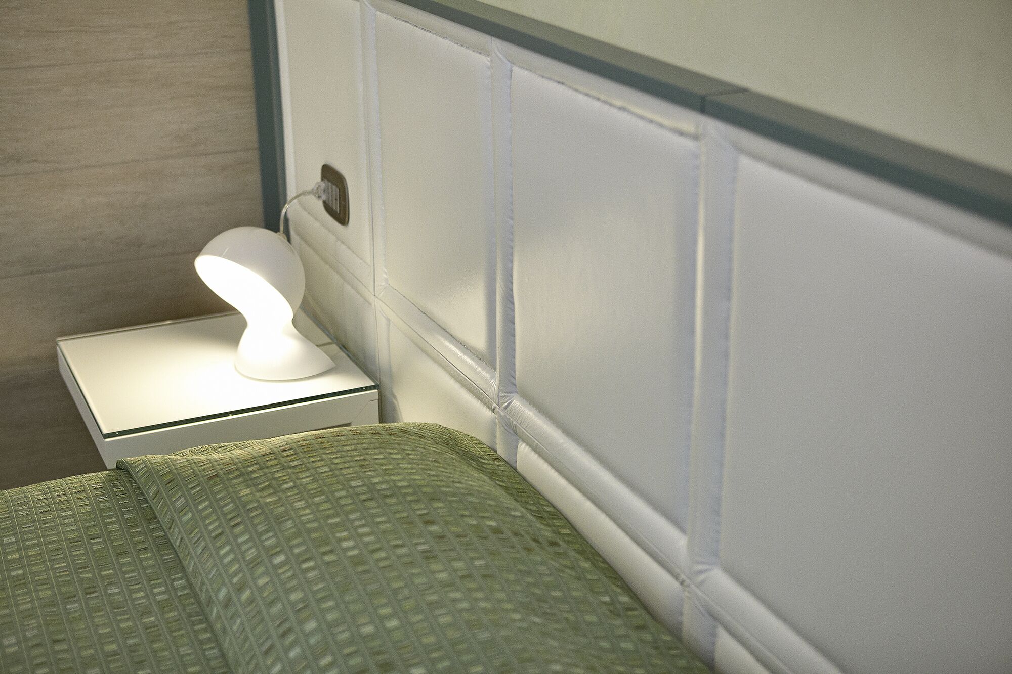 A neatly made bed with a green textured blanket, a white bedside lamp, and a white nightstand with a power outlet.