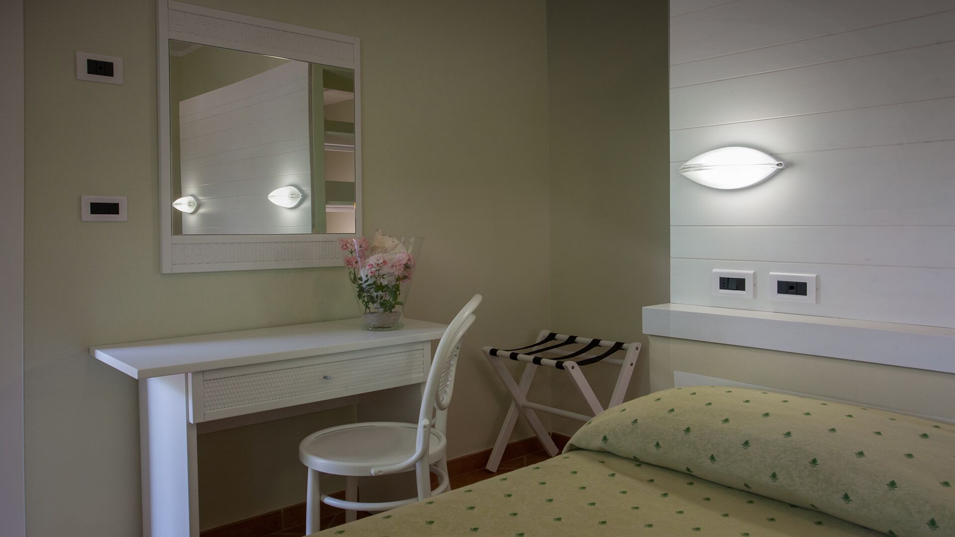 A bedroom with a bed, a desk, a chair, a mirror, and bedside lighting.
