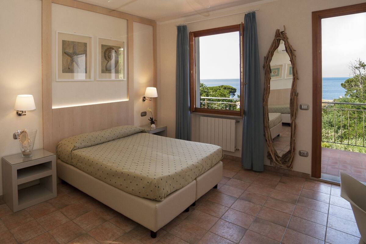 Ein Schlafzimmer mit einem Bett, zwei Nachttischlampen, einem Nachttisch, einem Fenster mit Vorhängen, Blick auf das Meer und einem Spiegel an der Wand.