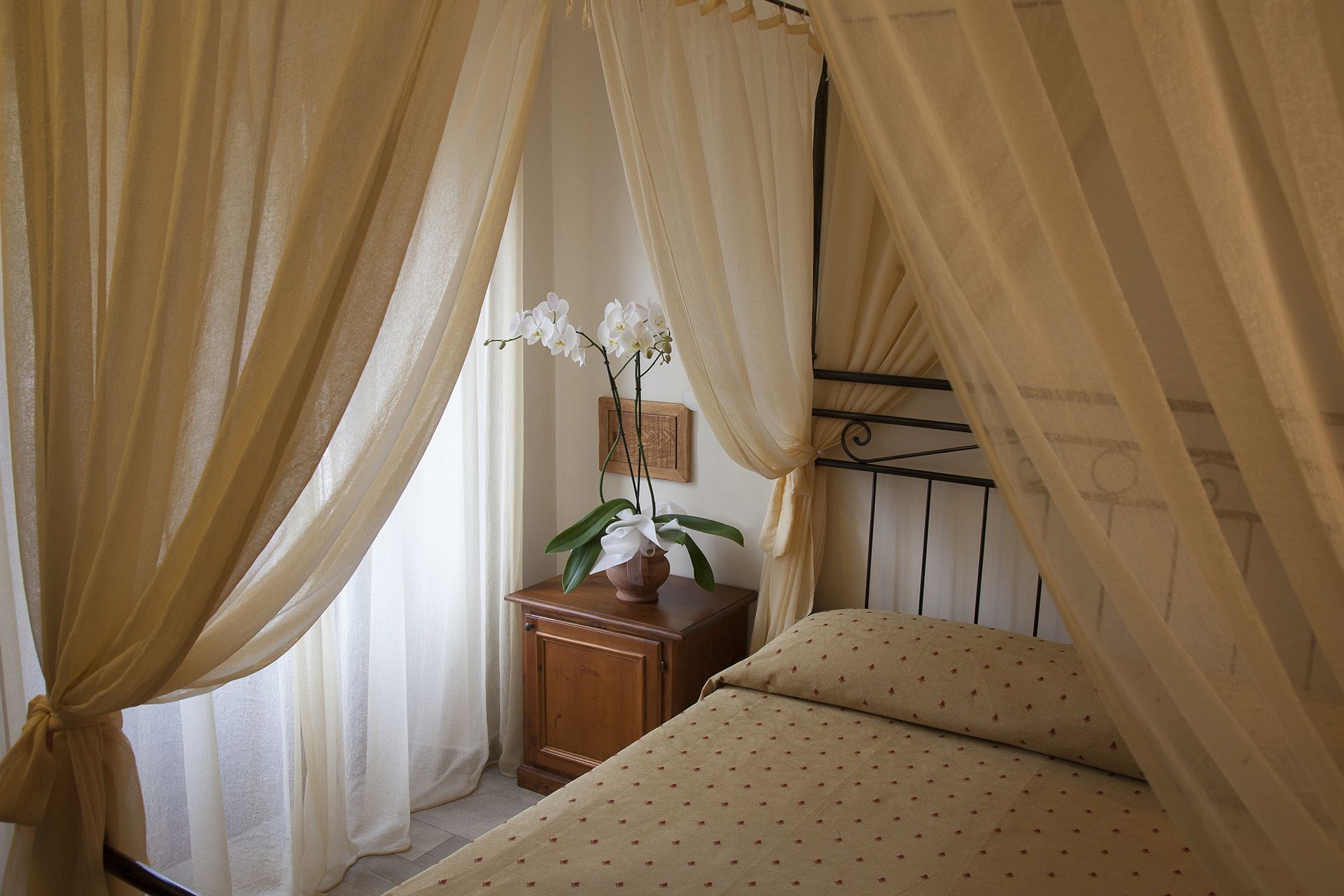 Un dormitorio con cama, mesita de noche, orquídea en maceta y cortinas traslúcidas.