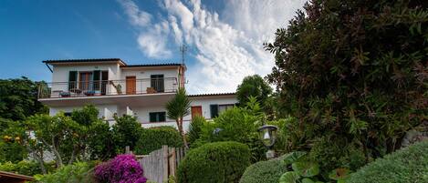 Una casa de dos pisos con balcón, rodeada de exuberante follaje y plantas florecientes.