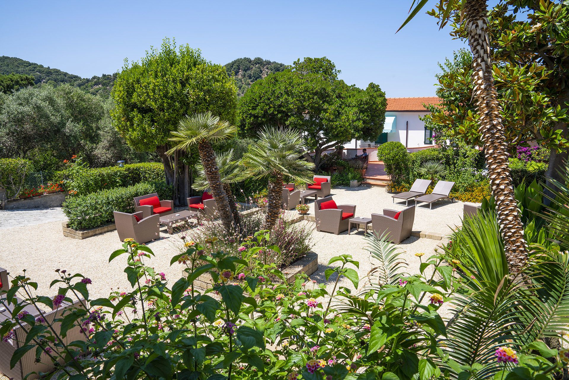 Um jardim com área de estar ao ar livre, um edifício com telhado de telhas vermelhas e vegetação exuberante.