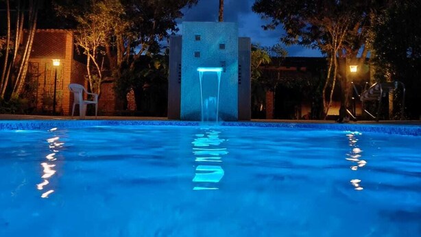A pool with a water feature in the center, surrounded by palm trees and a building with a lighted entrance. - Pousada Barcelos (São Roque de Minas)