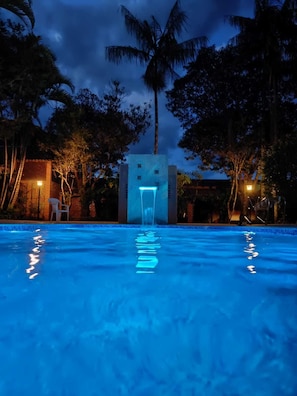 Piscina con una fuente en el centro, rodeada de palmeras y un edificio con entrada iluminada.