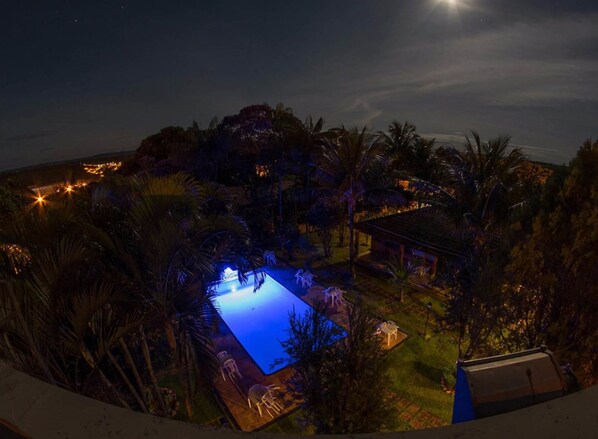 A nighttime view of a pool area surrounded by palm trees and a small building. - Pousada Barcelos (São Roque de Minas)