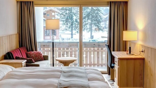 A hotel room with a bed, a sofa with pillows, a small table, a lamp, and a view of a snowy landscape through large windows. - City Hotel Haupthaus and Nebenhaus (Taesch)