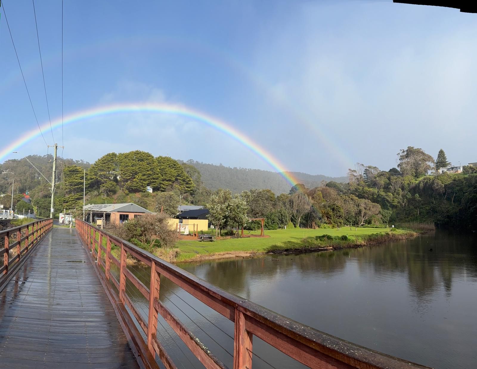 A wooden bridge with a metal railing over a calm body of water, surrounded by greenery and hills.