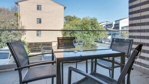 A balcony with a glass table and four chairs, overlooking a residential area with a brick building and parked cars.