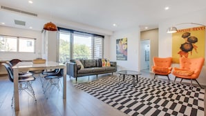 A modern living room with a black and white checkered rug, a sofa, two orange armchairs, a dining table with chairs, and a large window.