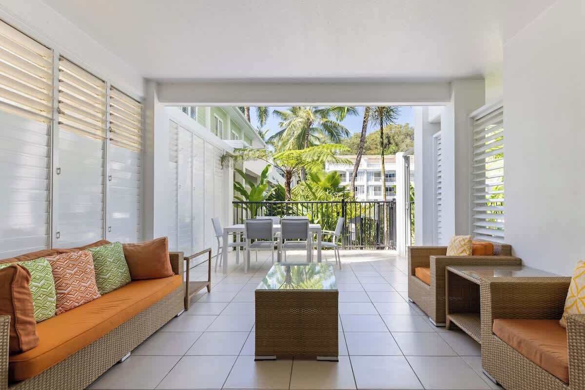 Un espace intérieur moderne comprenant un canapé, des fauteuils et une table basse. Un balcon offre une vue sur les palmiers et les immeubles.