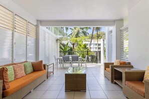 Un ambiente interno moderno con un divano, poltrone e un tavolino. È presente un balcone con vista su palme e edifici.