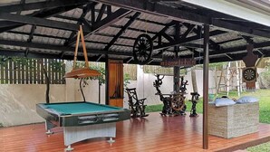 Une table de billard placée sous une zone abritée, avec un aménagement de style rustique, comprenant un luminaire suspendu et une horloge.