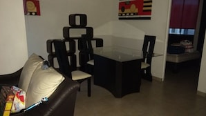 Un área de comedor con una mesa de vidrio y sillas negras, un sofá de cuero con un cojín, y una bolsa de papas sobre el sofá.
