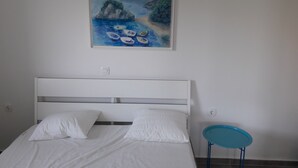 Un dormitorio con una cama, almohadas blancas, una silla azul y un cuadro de una escena costera.