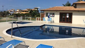 A swimming pool with lounge chairs, a small building with a tiled roof, and a view of houses in the background. - Beautiful house in the best gated community in Manguinhos. (Armação dos Búzios)