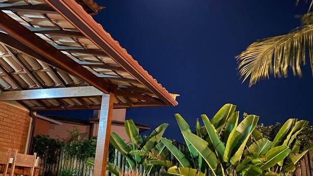 A nighttime view of a covered patio area with a brick wall, a wooden pergola, and lush tropical plants. - Beautiful house in the best gated community in Manguinhos. (Armação dos Búzios)