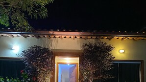 A well-lit entrance with a stone pathway leading to a wooden door, surrounded by lush greenery and flowering plants. - Beautiful house in the best gated community in Manguinhos. (Armação dos Búzios)
