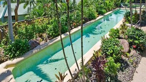 A rectangular swimming pool surrounded by lush greenery and palm trees, with a clear view of the ocean in the background. - Blue water Views 15, Beautiful Apartment on Hamilton Island (Hamilton Island)