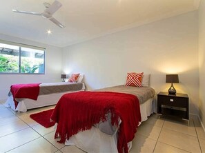Une chambre avec deux lits, un ventilateur de plafond, une table de chevet avec une lampe et une fenêtre donnant sur un paysage verdoyant.