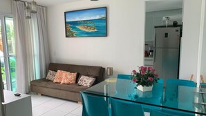 A modern living room with a glass dining table, teal chairs, a sofa with patterned cushions, a painting of a tropical scene, and a kitchen area with a stainless steel refrigerator.