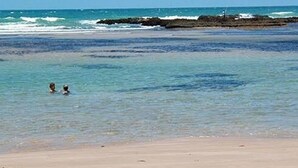 A beach with clear water, two people swimming, and a rocky outcrop in the distance.