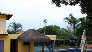 A swimming pool area with a thatched umbrella, a poolside chair, and a view of a building with a tiled roof. - Casa 4 suites a 200m da Rua das Pedras (Búzios)