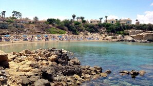 A coastal beach with rocky foreground, clear water, and houses in the background. - Kyklades Resort DPSBF04 -Colorful One Bed Apartment- WIFI-Communal Pool-Gym-Spa (Pralimni)