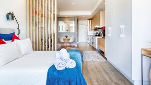 Una habitación de hotel moderna con una cama, una manta azul y toallas. Se observa una división de madera, un escritorio y una kitchenette al fondo.