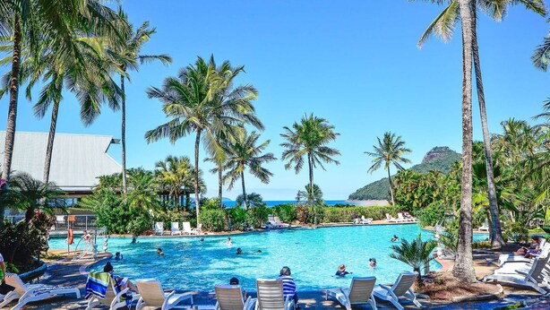 Ein Poolbereich mit Liegestühlen, Palmen und Blick aufs Meer.