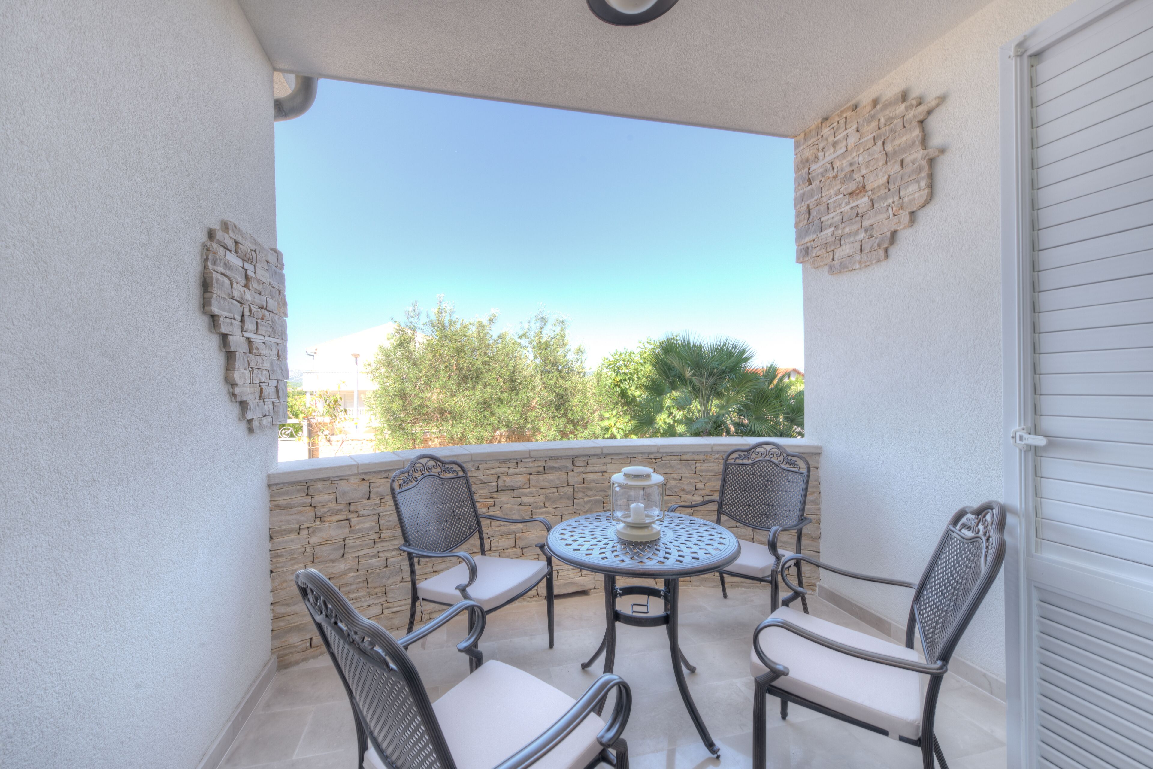 A balcony with a round table and four chairs, a stone wall, and a view of trees and buildings.