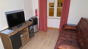 Un salon avec un téléviseur posé sur un meuble en bois, un canapé, une petite poubelle et une fenêtre avec des rideaux.