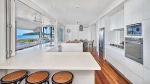 Cocina moderna con una isla grande, muebles blancos y electrodomésticos de acero inoxidable. Cuenta con un comedor con mesa y sillas, y vistas al mar a través de amplios ventanales.