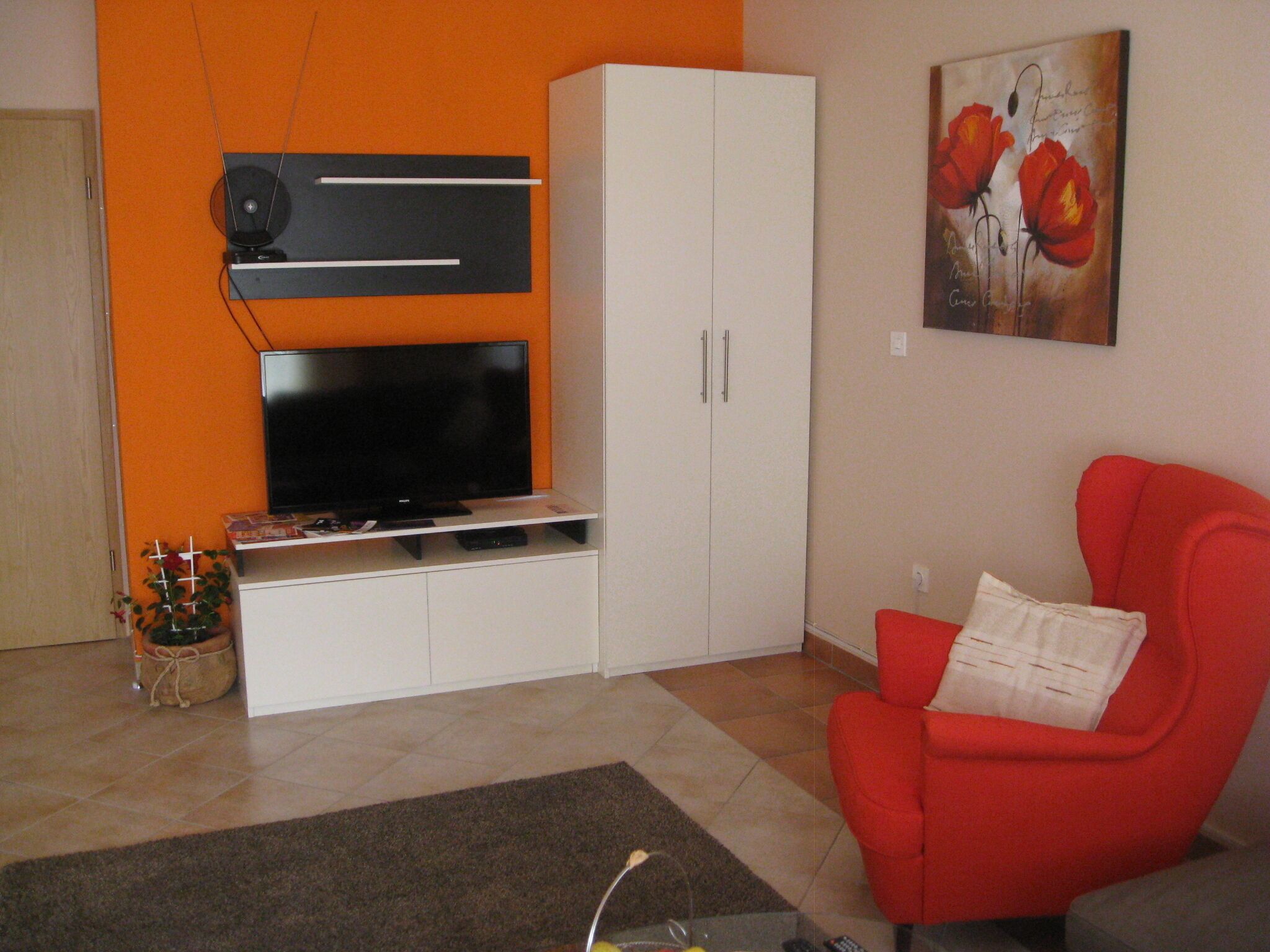 Un salon doté d’un téléviseur à écran plat, de meubles blancs, d’un fauteuil rouge et d’un tableau représentant des fleurs rouges accroché au mur.