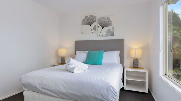 A neatly made bed with white linens and a folded towel, two bedside lamps, a grey headboard, and two wall-mounted artworks depicting abstract tree designs.
