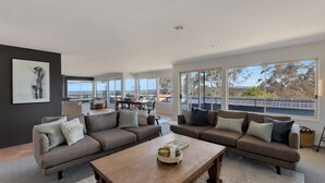 Amplia sala de estar con dos sofás, una mesa de centro y un comedor con mesa y sillas. Ventanas grandes ofrecen vistas al exterior.