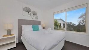 A neatly made bed with white linens and a teal pillow, a bedside table with a lamp, and a large window with a view of a house and trees.