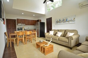 A modern kitchen with wooden cabinets and a black refrigerator, a dining area with wooden chairs and a table, and a living room with a beige sofa and a painting on the wall.