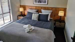 A neatly made bed with a grey and white duvet, two pillows, and a rolled white towel. There are two bedside tables with lamps, a framed picture on the wall, and a window with blinds.