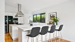 A modern kitchen with a central island, black bar stools, a large window, and a stainless steel range hood. - Pet Friendly - Miss Maude Mt Martha with pool and spa (Mount Martha)