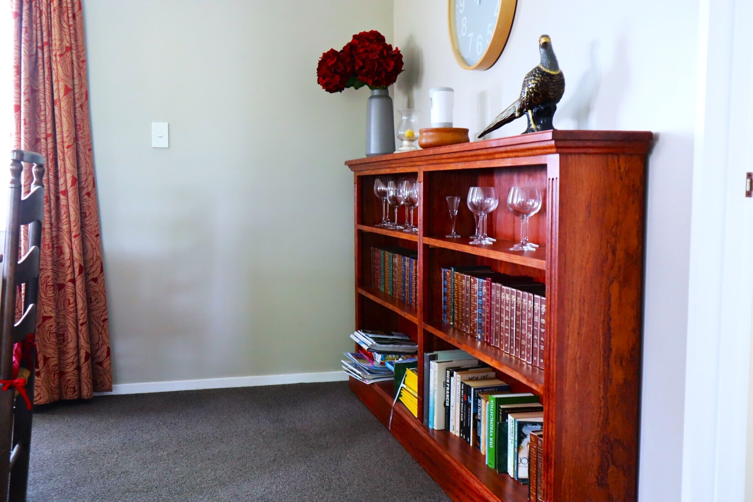 Une pièce avec une bibliothèque en bois remplie de livres et de verres à vin, un vase avec des fleurs rouges et une horloge murale.