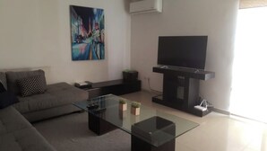 Uma sala de estar moderna com um sofá cinzento, uma mesa de centro de vidro, uma TV de tela plana sobre um suporte preto e um quadro na parede.