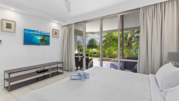 Quarto de hotel com uma cama grande, televisão de tela plana fixada na parede, ventilador de teto e vista para o exterior através de grandes portas de correr de vidro.