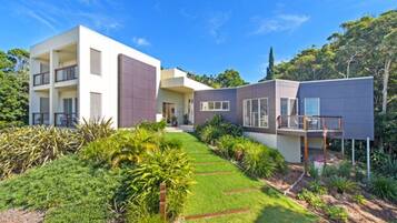 Une maison moderne à deux étages, dotée d’un toit plat, de grandes fenêtres et d’un jardin bien entretenu, agrémenté de diverses plantes et d’un sentier.
