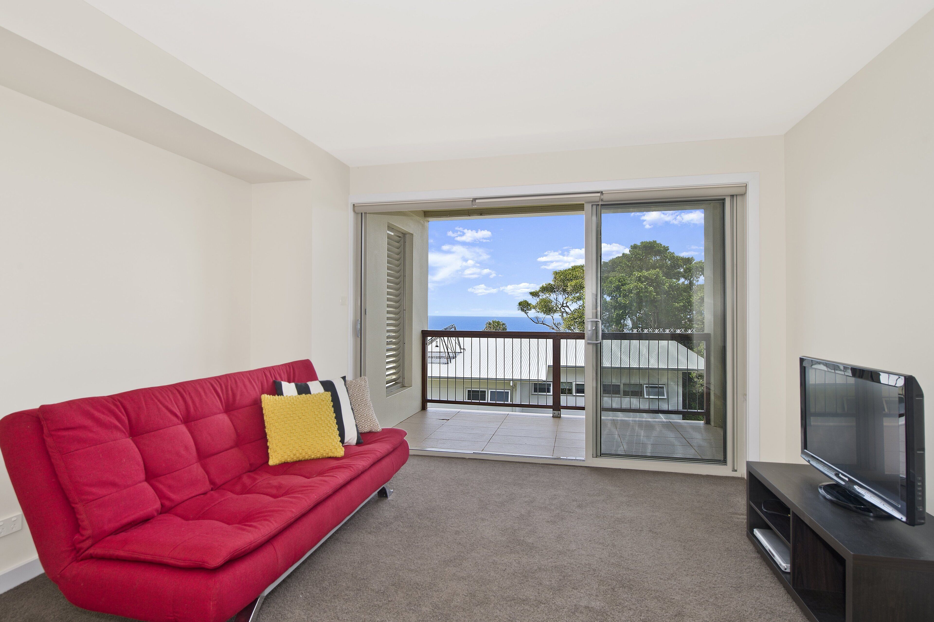 A modern living room with a red sofa, a flat-screen TV, and a balcony with a view of the sea.
