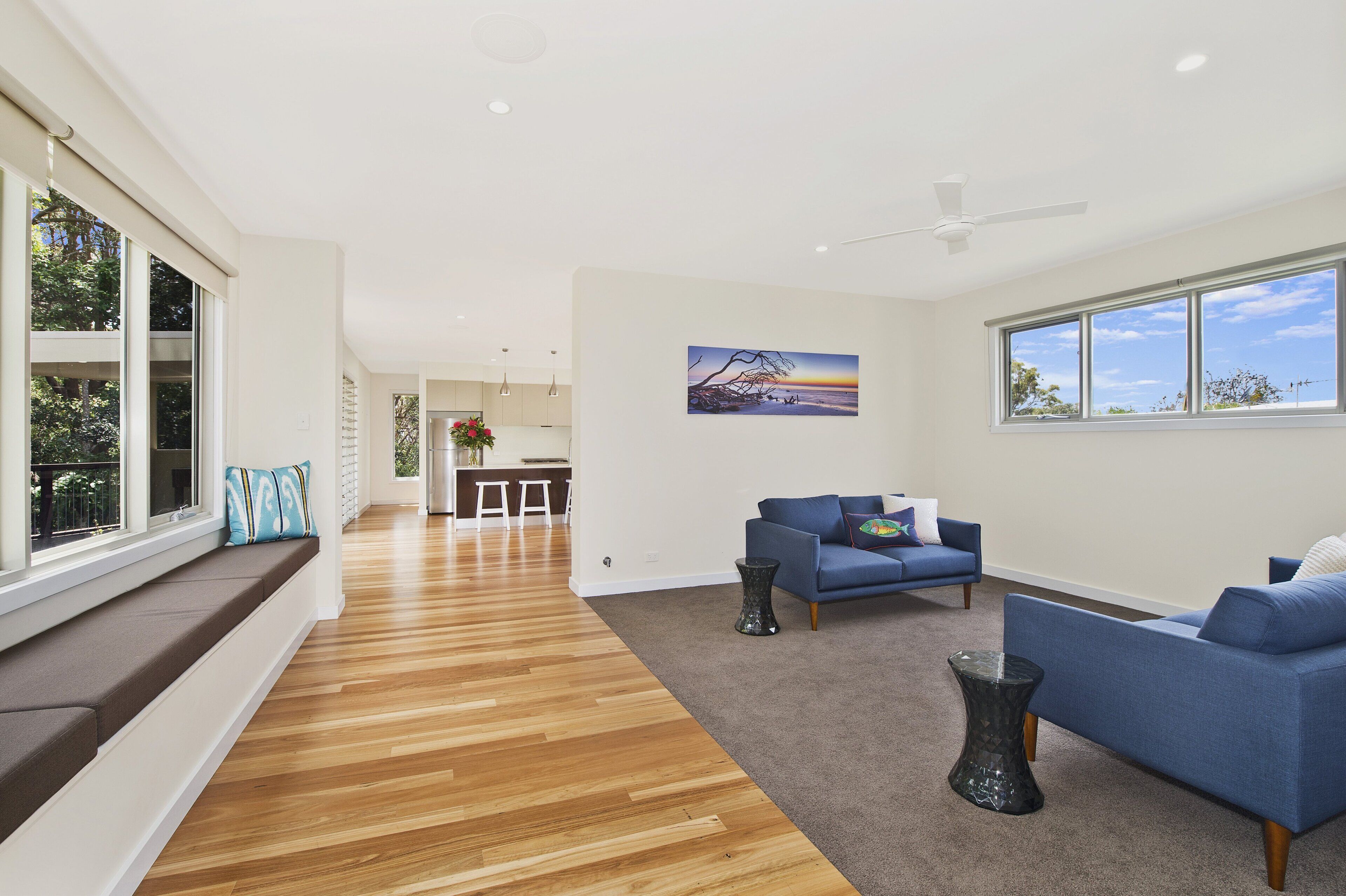 A spacious living area with a wooden floor, blue sofas, a dining area, and large windows offering views of the outdoors.