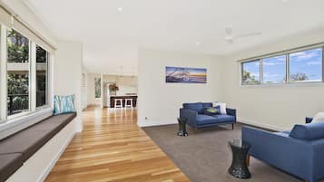 A spacious living area with a wooden floor, blue sofas, a dining area, and large windows offering views of the outdoors.