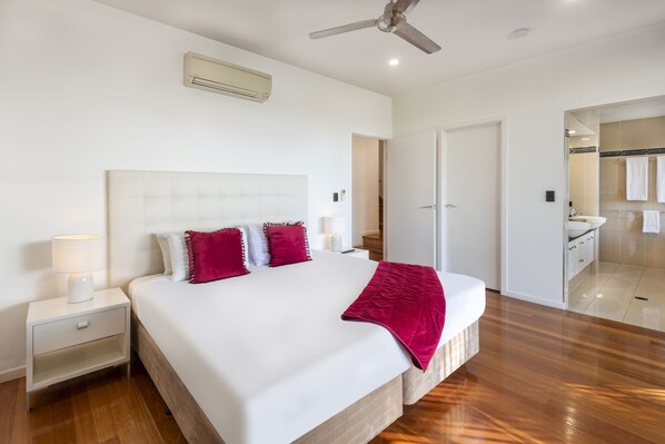 A modern bedroom with a large bed, white walls, a ceiling fan, and a bathroom visible through an open door.