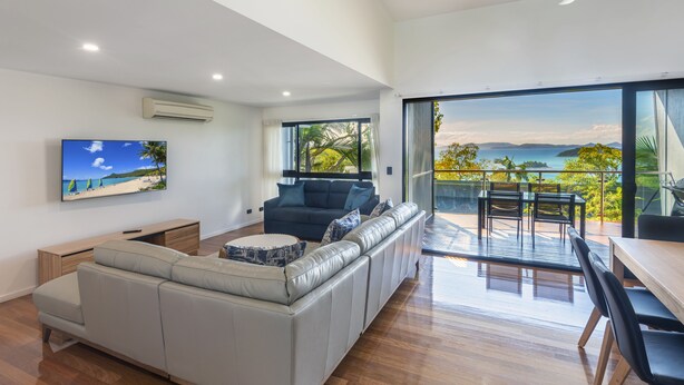 A modern living room with a large sofa, a dining area with a table and chairs, and a balcony with a view of the sea. - Pinnacle 2, Seaview Apartment on Hamilton Island (Hamilton Island)
