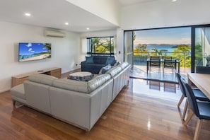 A modern living room with a large sofa, a dining area with a table and chairs, and a balcony with a view of the sea.