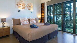 A modern bedroom with a large bed, a nightstand, a wall-mounted shelf, and a large window with a view of trees.